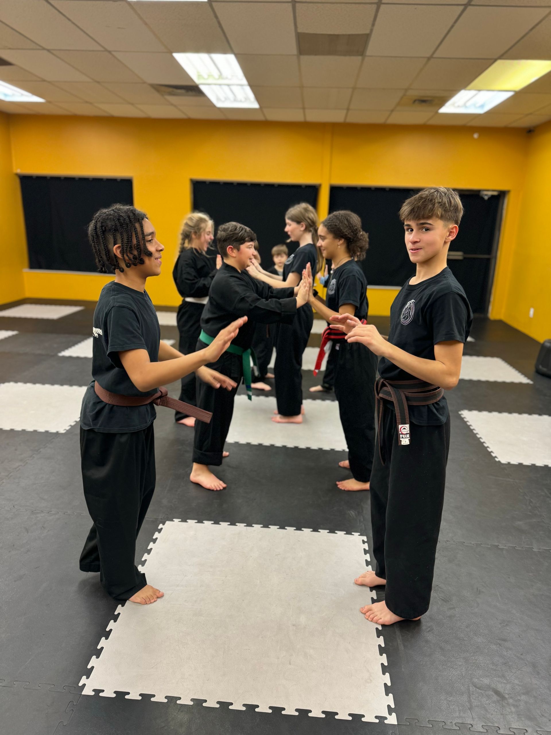 Students in black uniforms practice hand-to-hand drills in a martial arts studio with black and white floor mats.