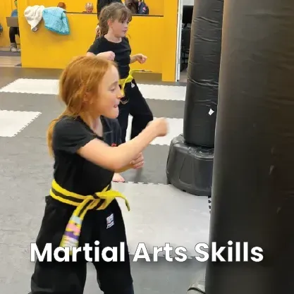 Two students in black uniforms and yellow belts practice punching a free-standing heavy bag in a martial arts studio.