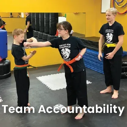 Three students in black martial arts uniforms practice a fist-bump greeting in a studio with a yellow wall and floor mats.