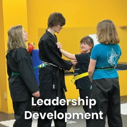 A martial arts instructor assists a younger student in a black uniform and yellow belt in a bright yellow training studio.