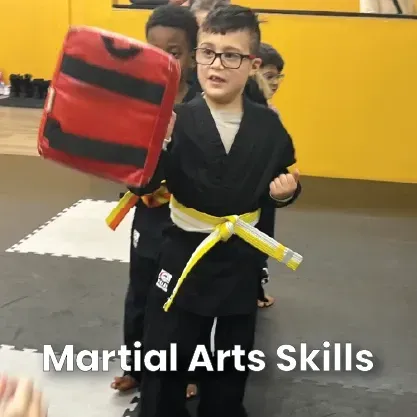 A child in a black martial arts uniform and yellow belt holding a red striking pad in a studio.
