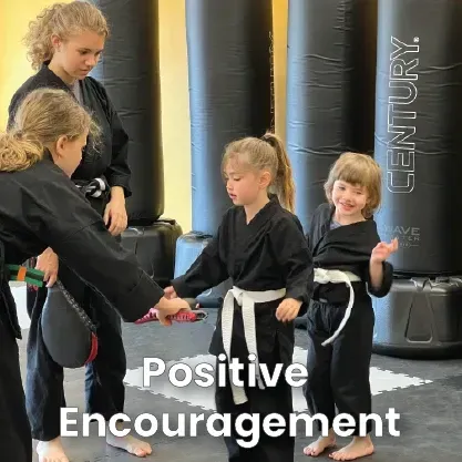 In a martial arts studio, an instructor helps a student with a practice strike while another student smiles nearby.