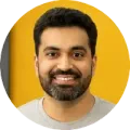 A smiling person with dark hair and a beard, wearing a grey crew-neck shirt against a vibrant yellow circular background.