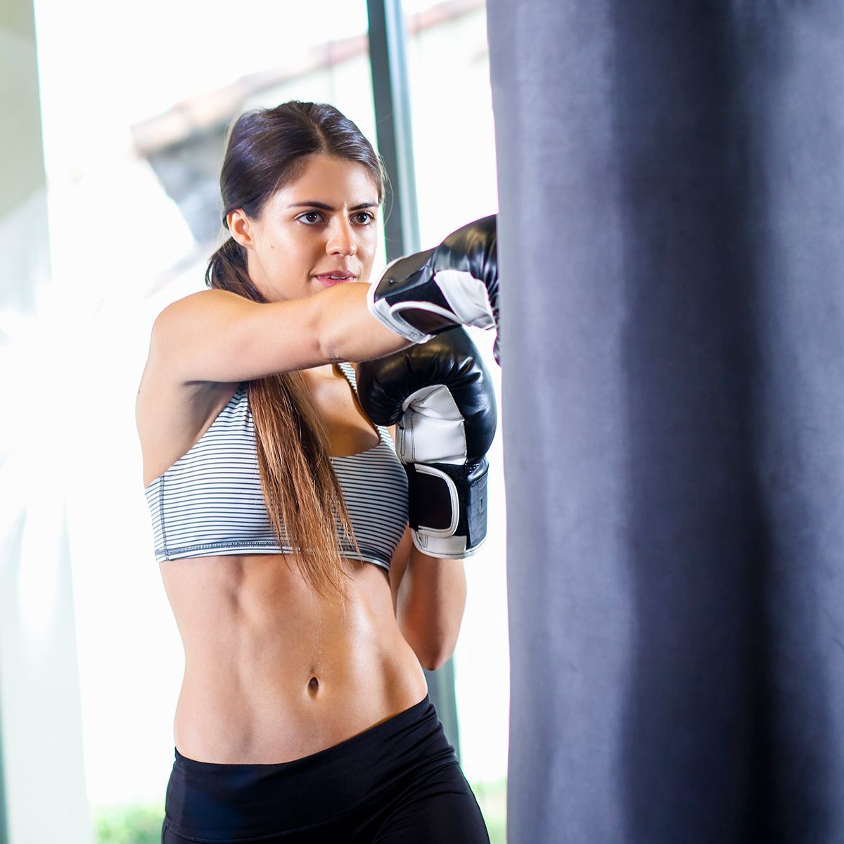 A person wearing boxing gloves strikes a heavy black punching bag in an indoor gym setting.