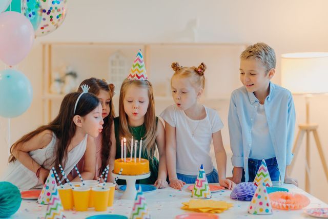 Four children lean over a small birthday cake with lit candles, preparing to blow them out at a decorated party table.