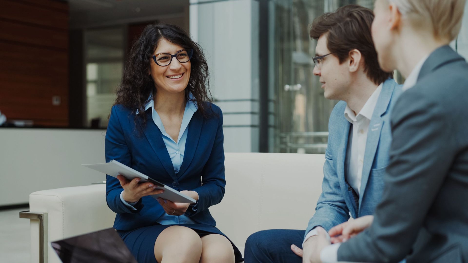 Three colleagues in business attire discussing a tablet in a modern office lounge.