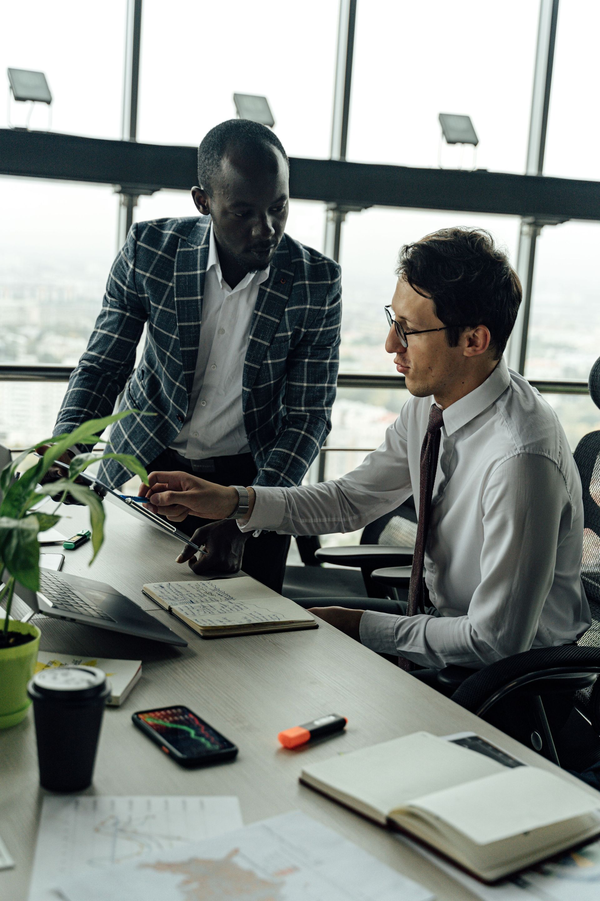 Two coworkers discuss work at a desk with laptop, notebook, and coffee in a bright office