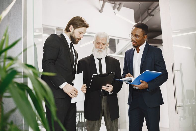 Three business people in suits discussing documents in a bright office hallway