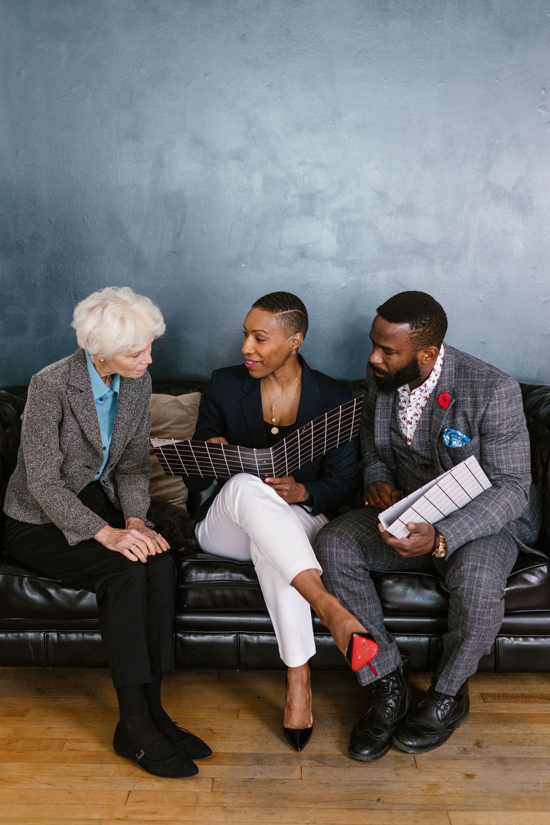 Three people seated on a black couch chatting in a room with a blue-gray wall and wooden floor.