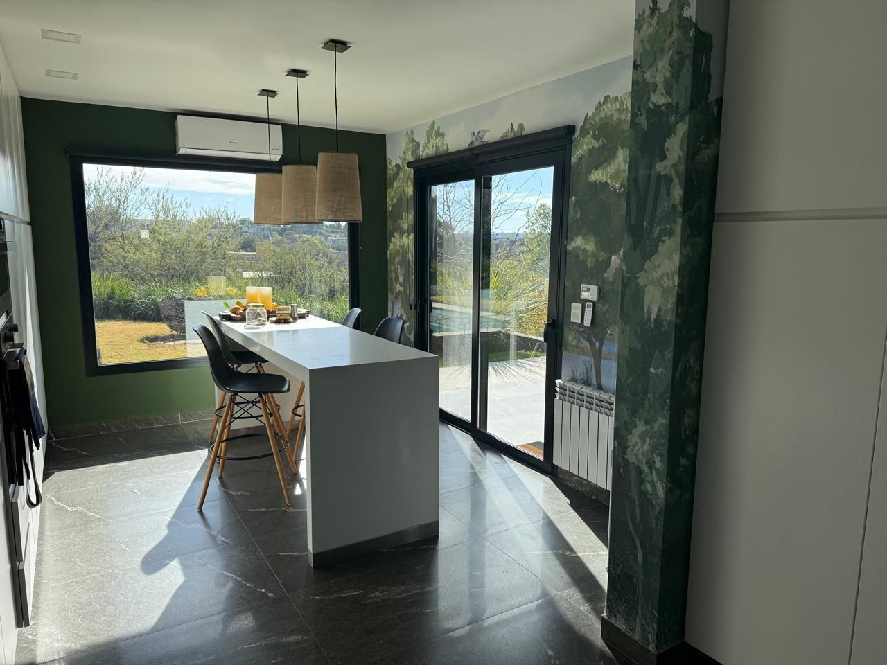 Cocina con isla y sillas, grandes ventanales con vistas a un paisaje verde, pared de acento verde y luces colgantes tejidas.