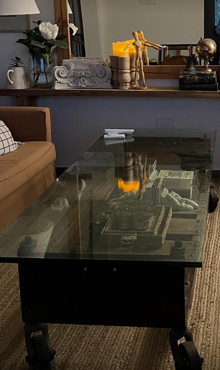 Una mesa de café con tapa de vidrio, marco oscuro y ruedas; una vela encendida en la parte superior refleja.