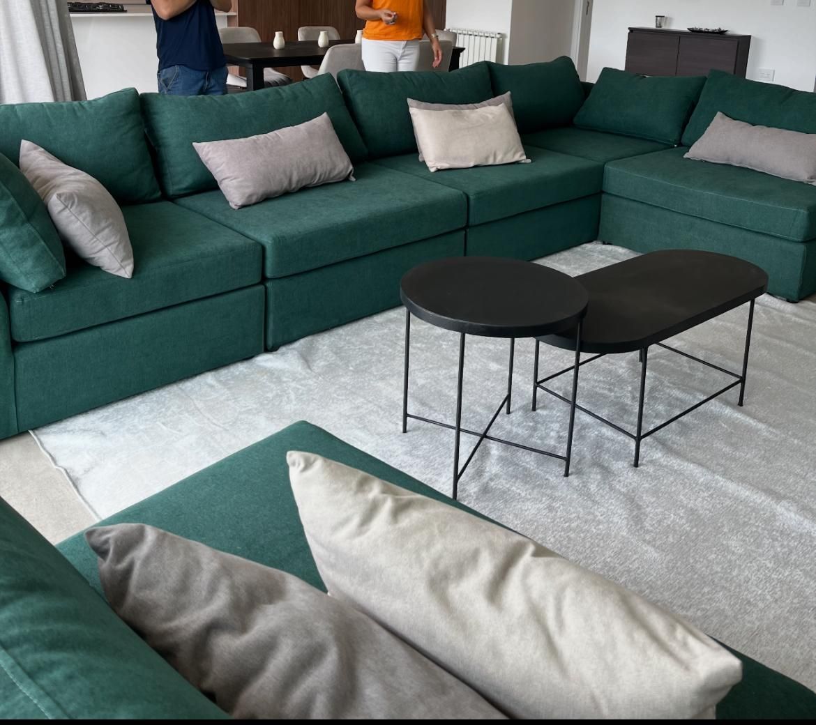 Sofá seccional verde esmeralda con almohadas grises y beige, dos mesas de café negras y una alfombra blanca.