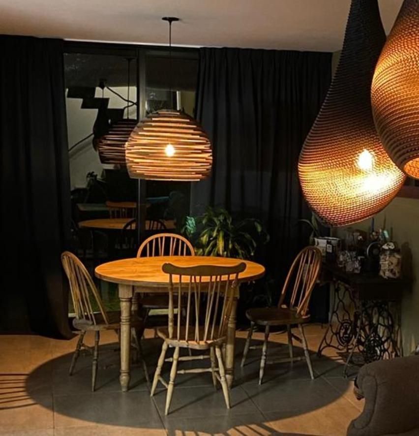 Comedor con mesa y sillas de madera y lámparas colgantes tejidas. Cortinas negras y vista al exterior.