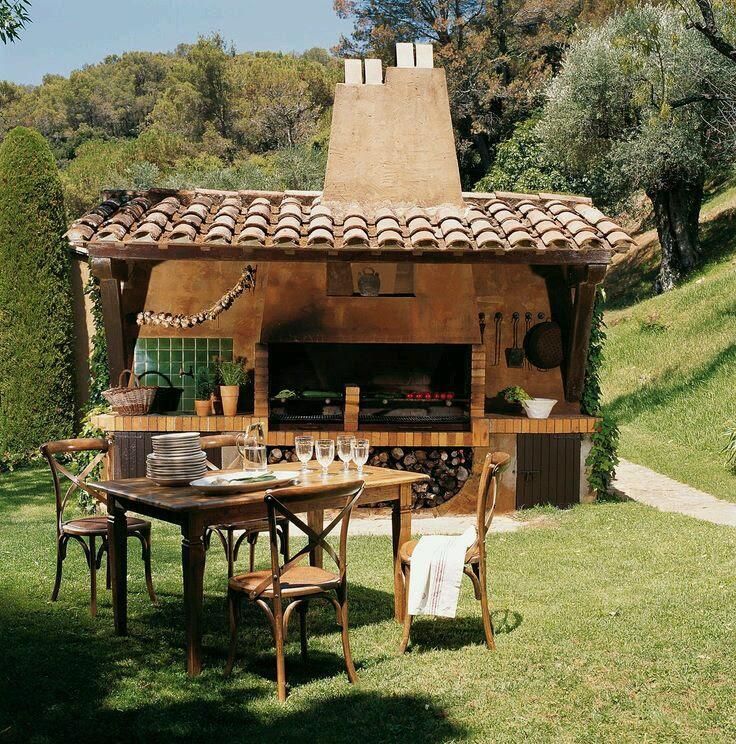Parrilla al aire libre con mesa y sillas sobre césped. Techo de tejas, chimenea de ladrillo y utensilios de cocina.