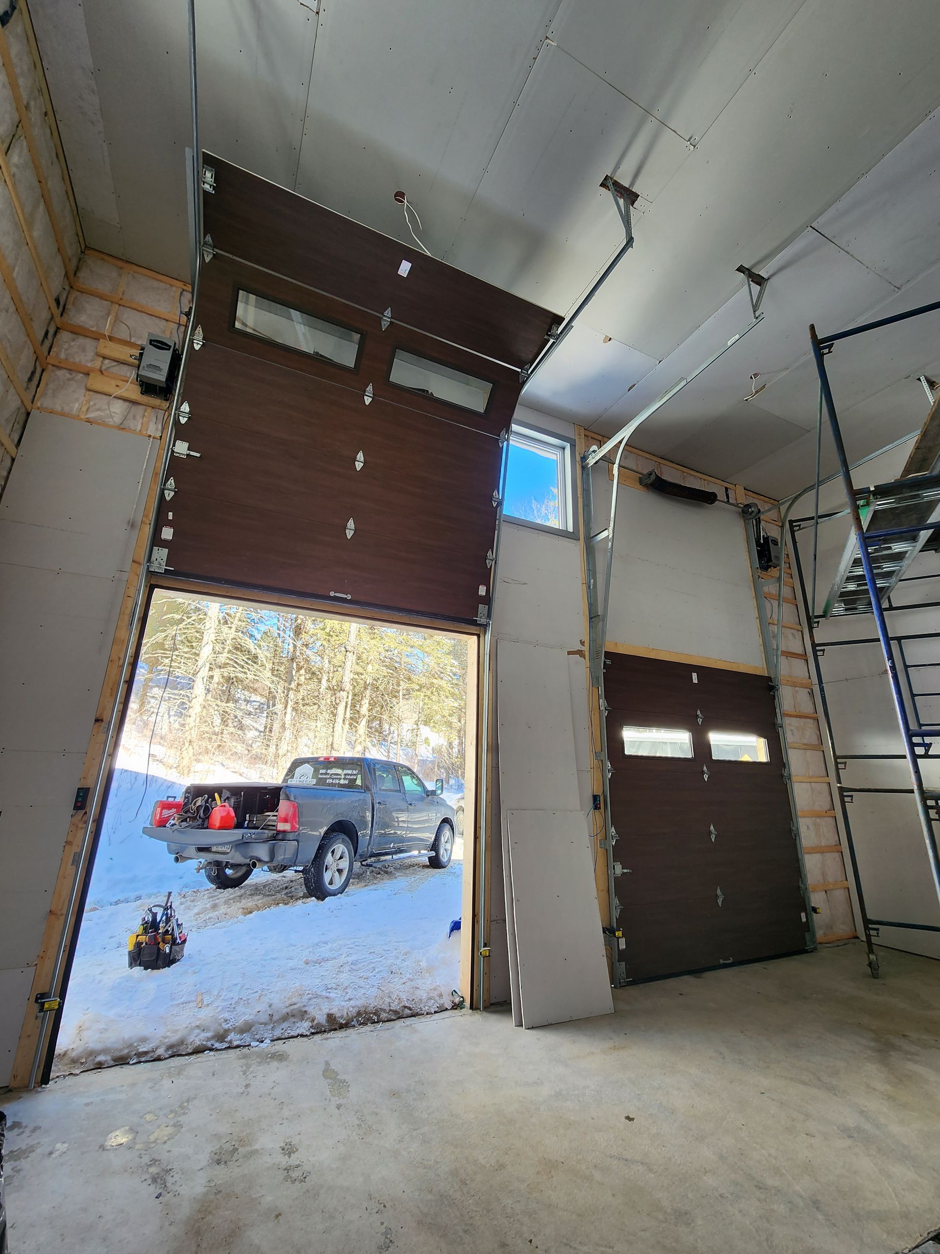 Maison moderne à deux étages avec revêtement en pierre et en bois, deux portes de garage noires et allée enneigée.
