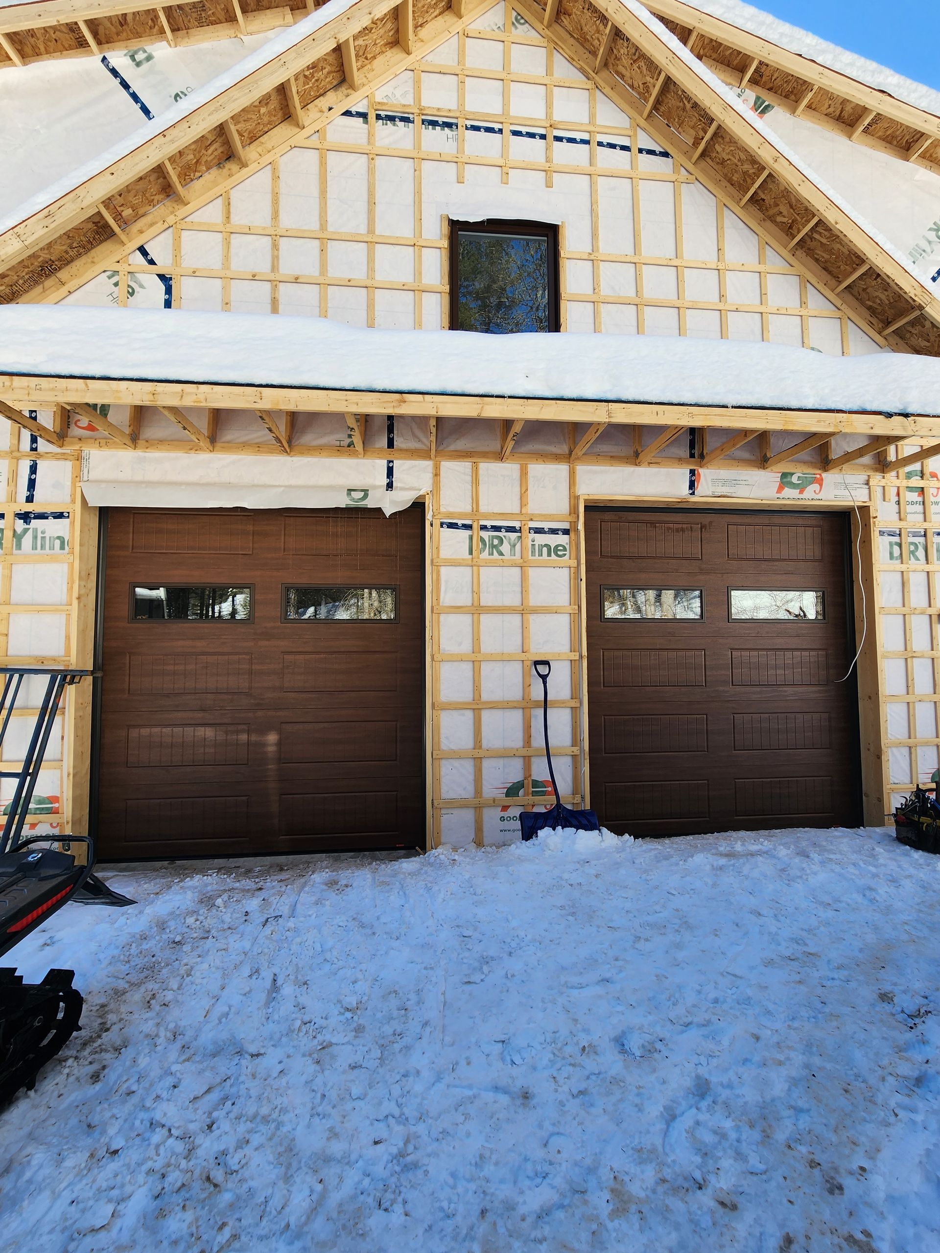 Maison moderne à deux étages avec revêtement en pierre et en bois, deux portes de garage noires et allée enneigée.