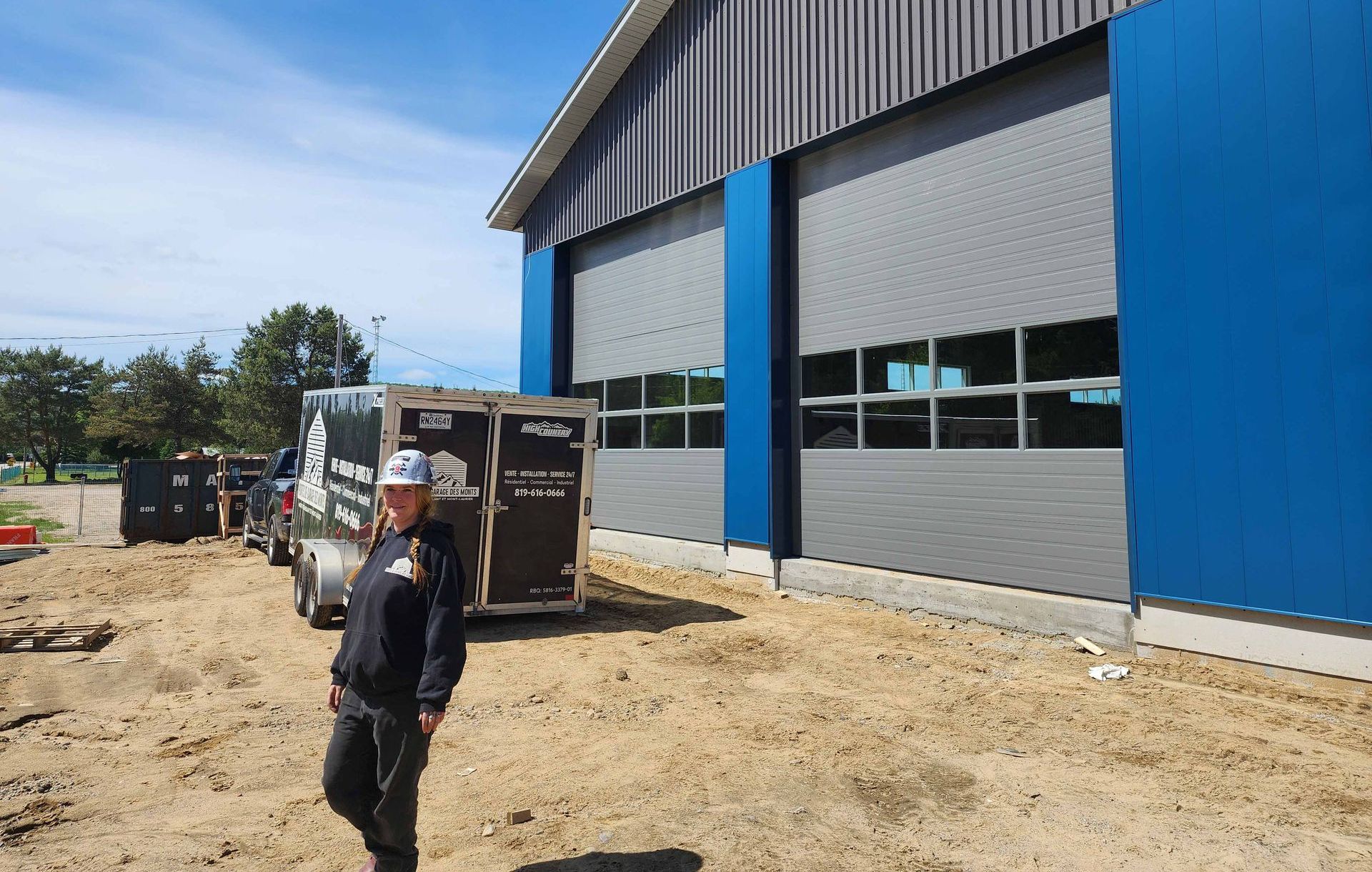 Une personne se tient près d'une remorque devant un bâtiment bleu avec des portes de garage grises.
