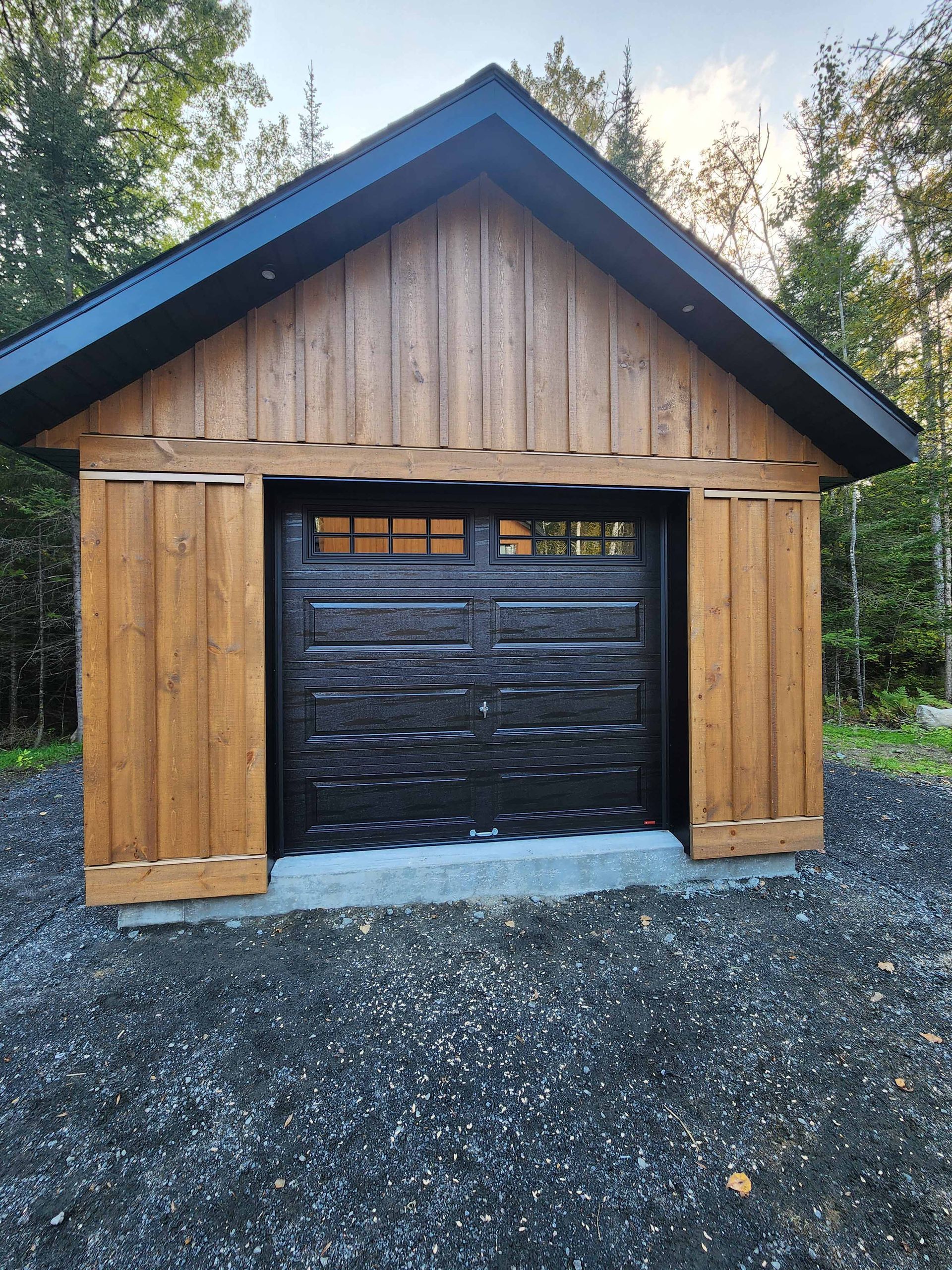 Garage marron avec porte et toit noirs, allée en gravier, fond de forêt.