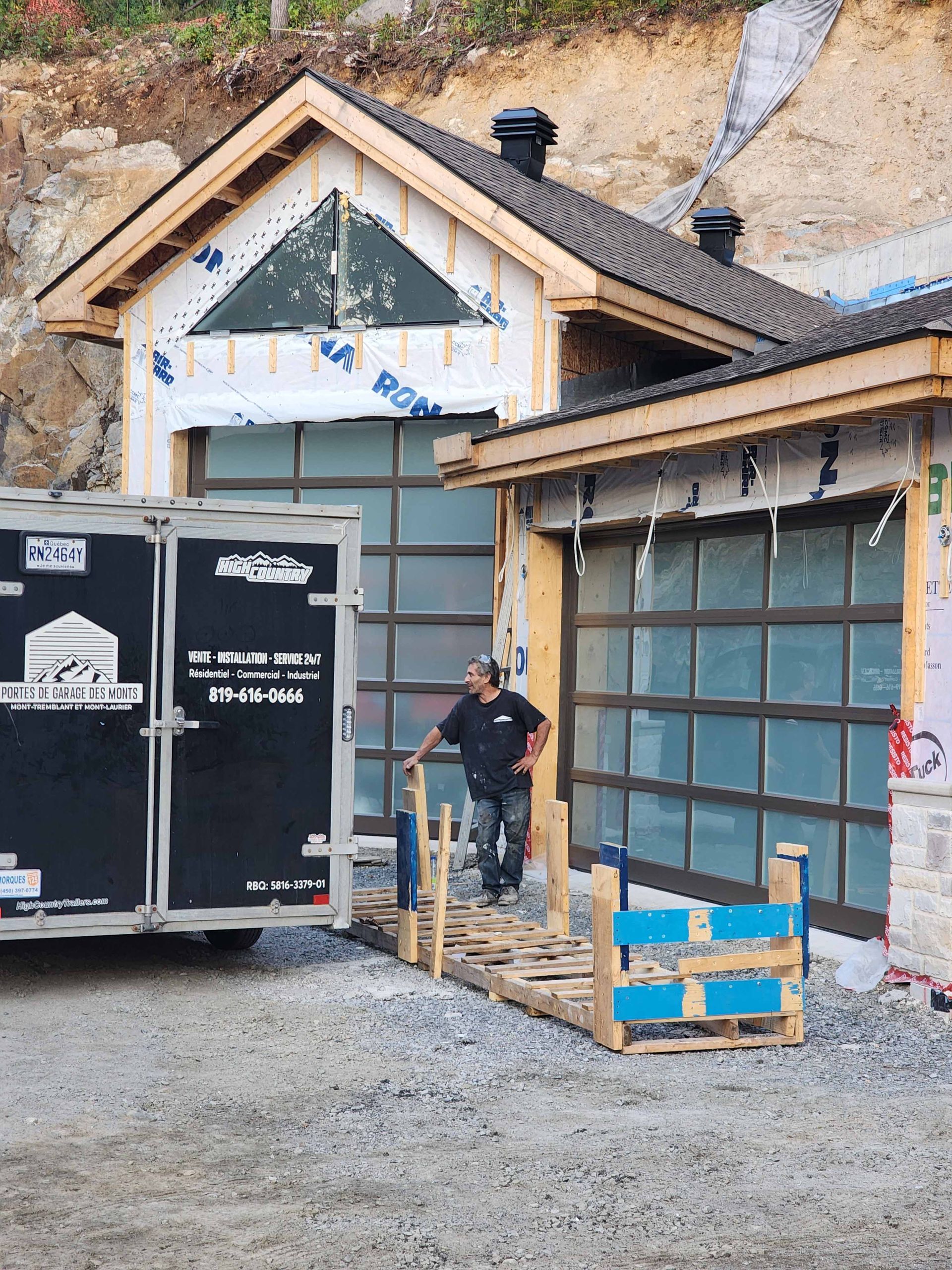 Un homme se tient sur une rampe devant des bâtiments aux portes de garage vitrées ; une remorque se trouve à proximité.