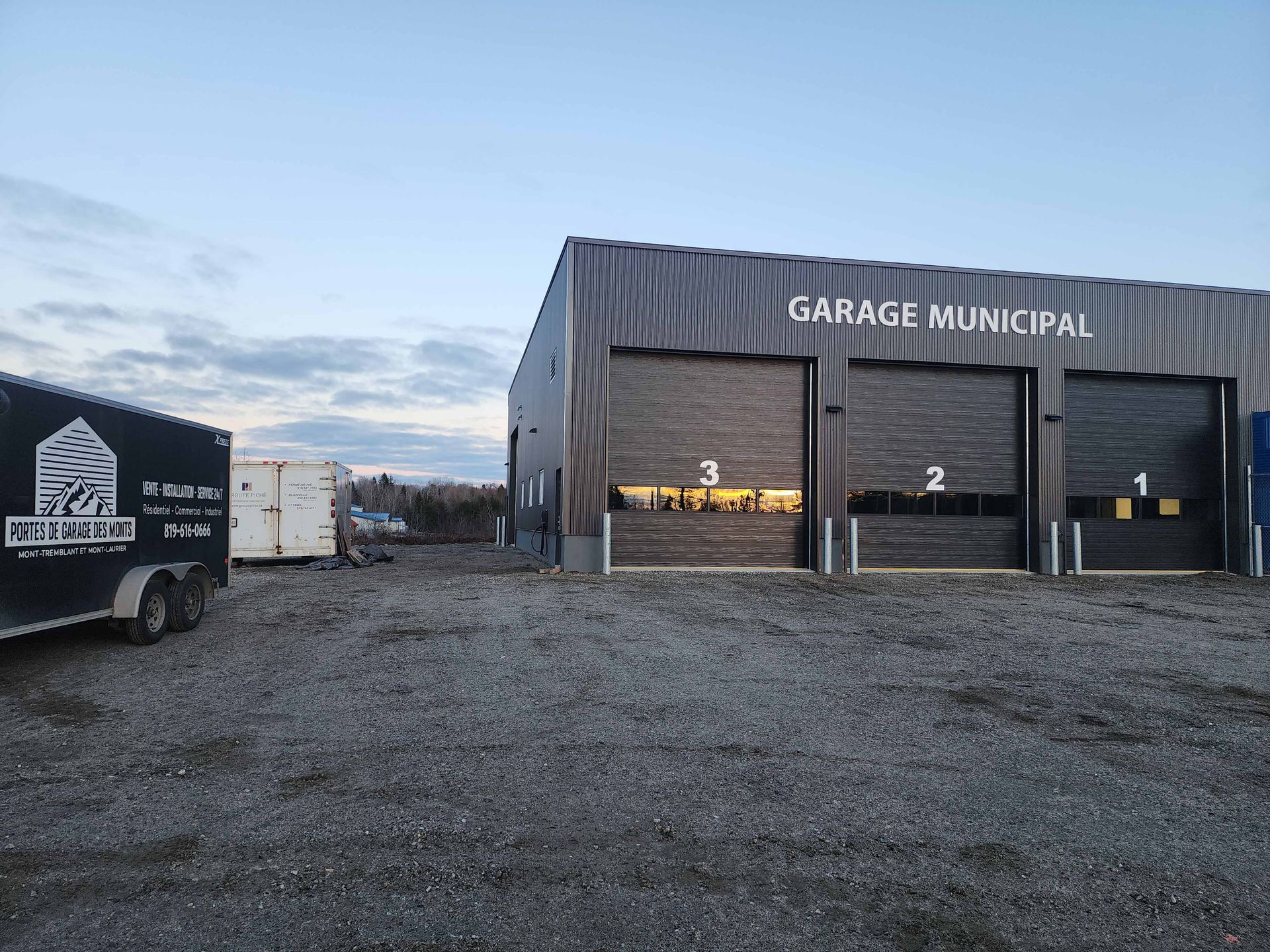 Garage municipal à trois emplacements numérotés. Extérieur noir, parking en gravier, ambiance crépusculaire.