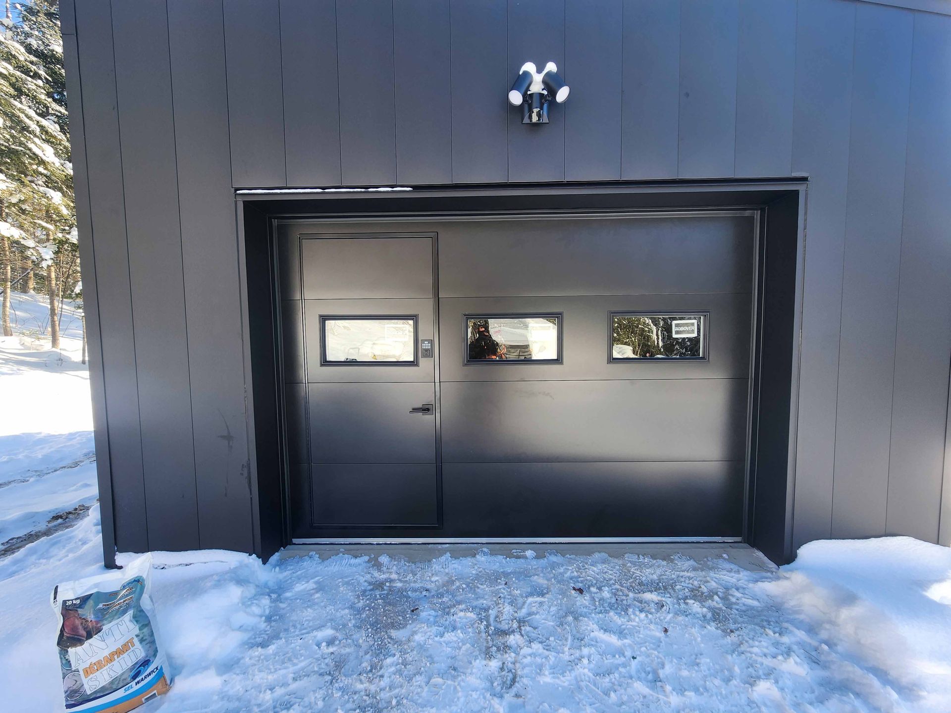 Porte de garage noire à trois fenêtres et éclairage extérieur, dans un décor enneigé.