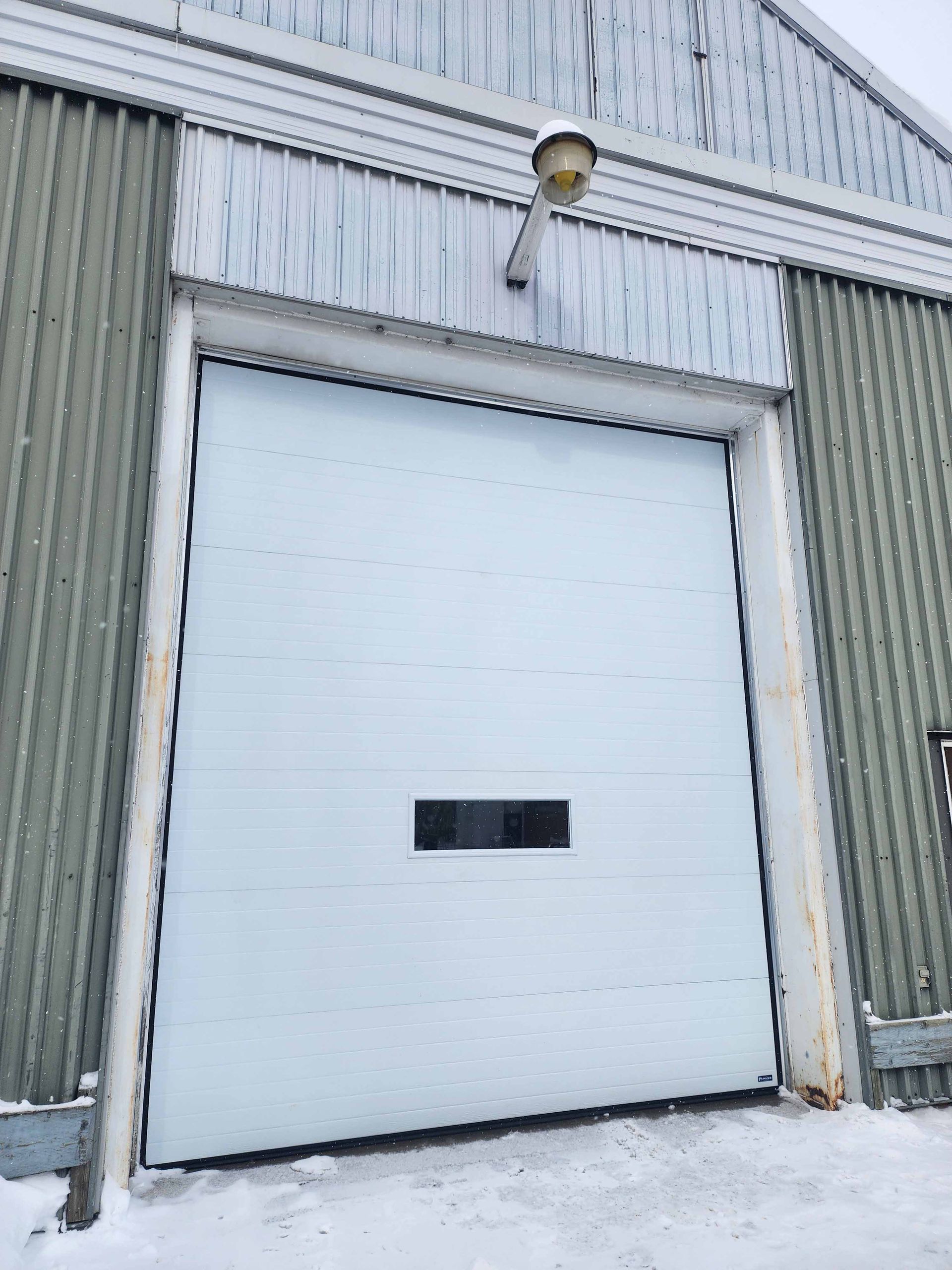 Porte de garage sectionnelle blanche dans un bâtiment en tôle ondulée, neige au sol.