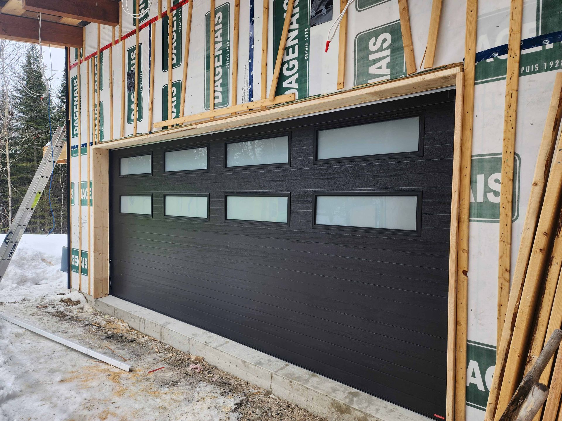 Porte de garage noire à vitres dépolies, installée dans un cadre en bois. Chantier, environnement extérieur.