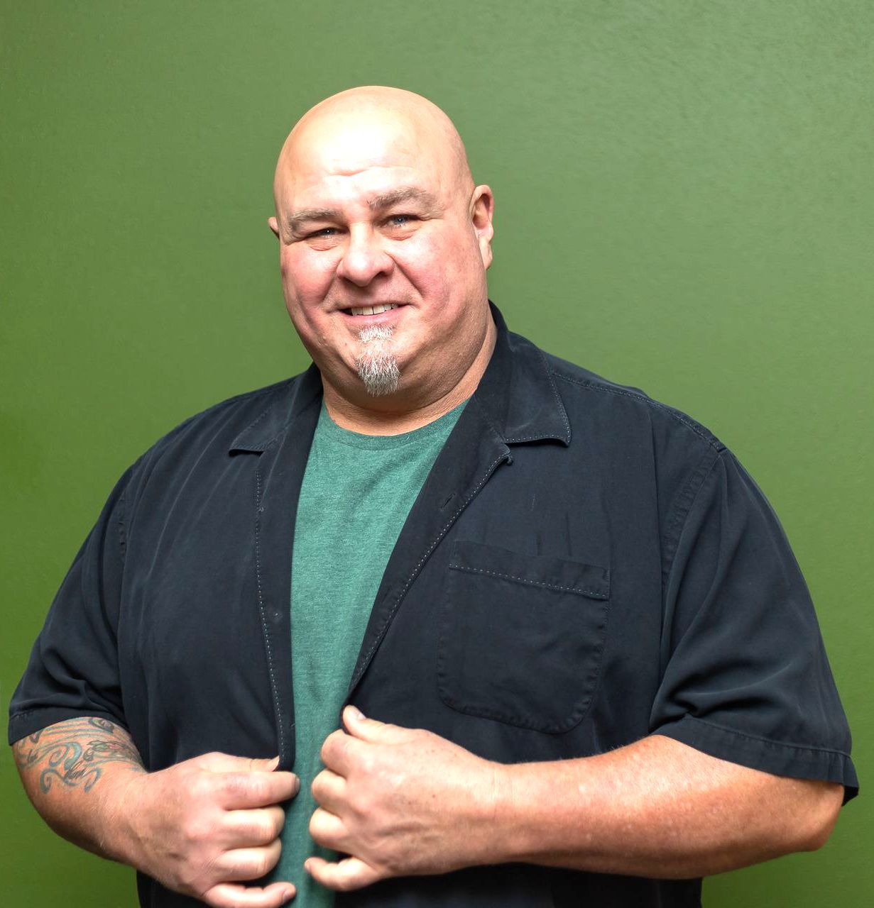 Smiling bald man in a black shirt over a green T-shirt, standing against a green background