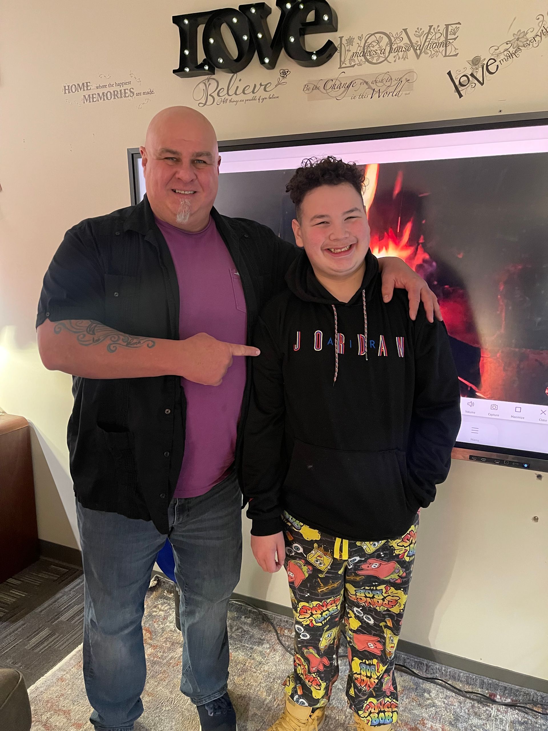 Two smiling people standing indoors by a wall-mounted TV and “love” wall decor, one pointing at the other.