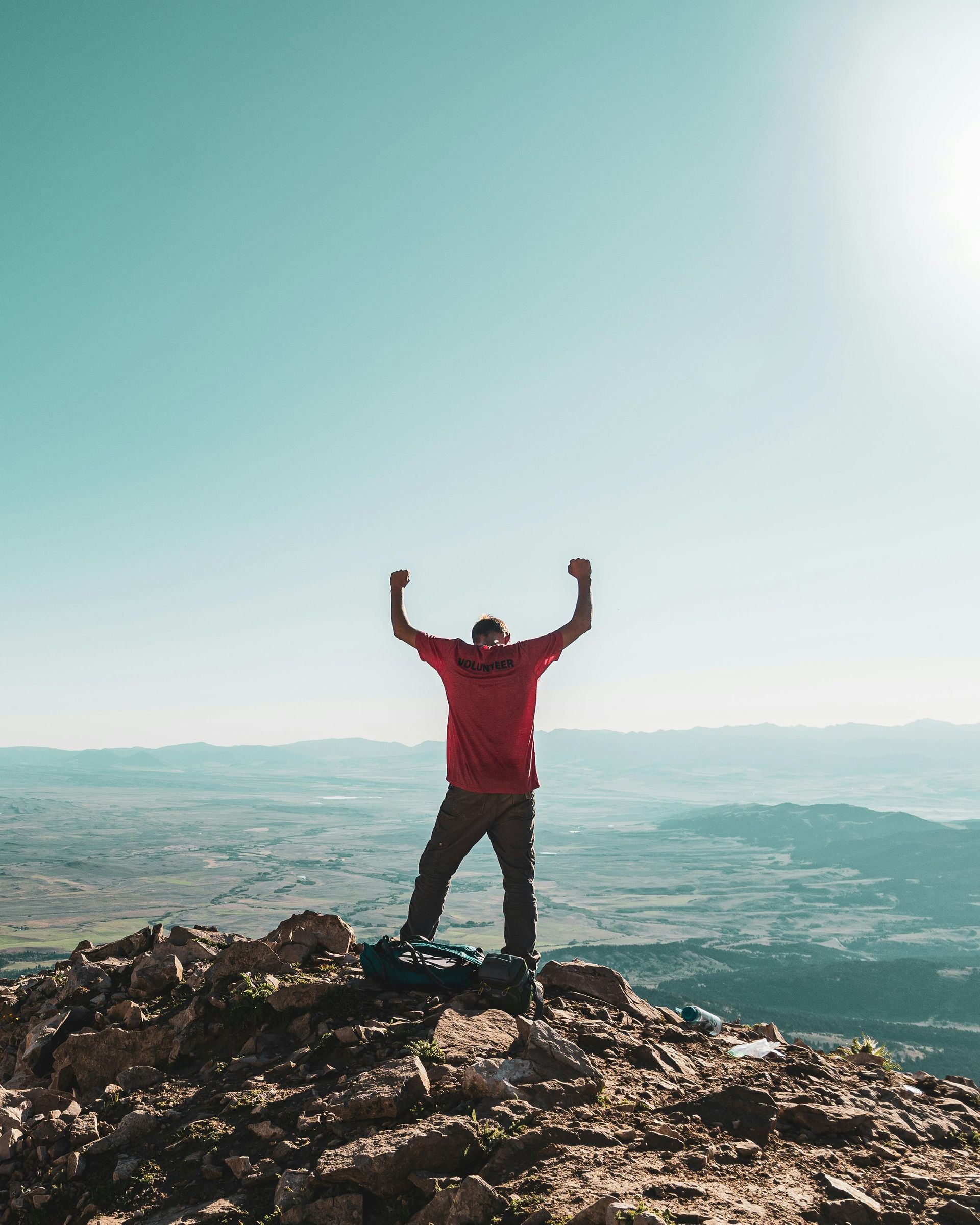 Person in red stands on a rocky summit with arms raised, overlooking a wide mountain landscape.