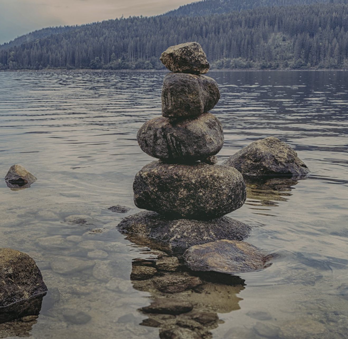 Stacked stones forming a cairn in a calm lake, with forested hills in the background.