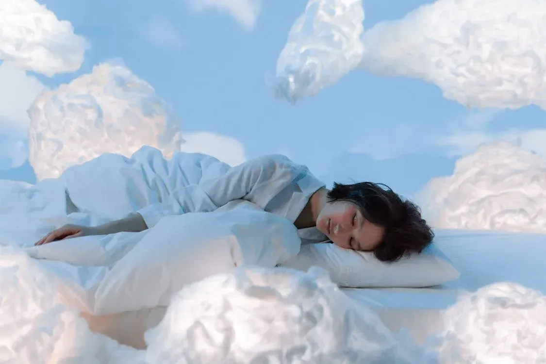 Person lying on white clouds under a blue sky, eyes closed, wearing a white outfit.
