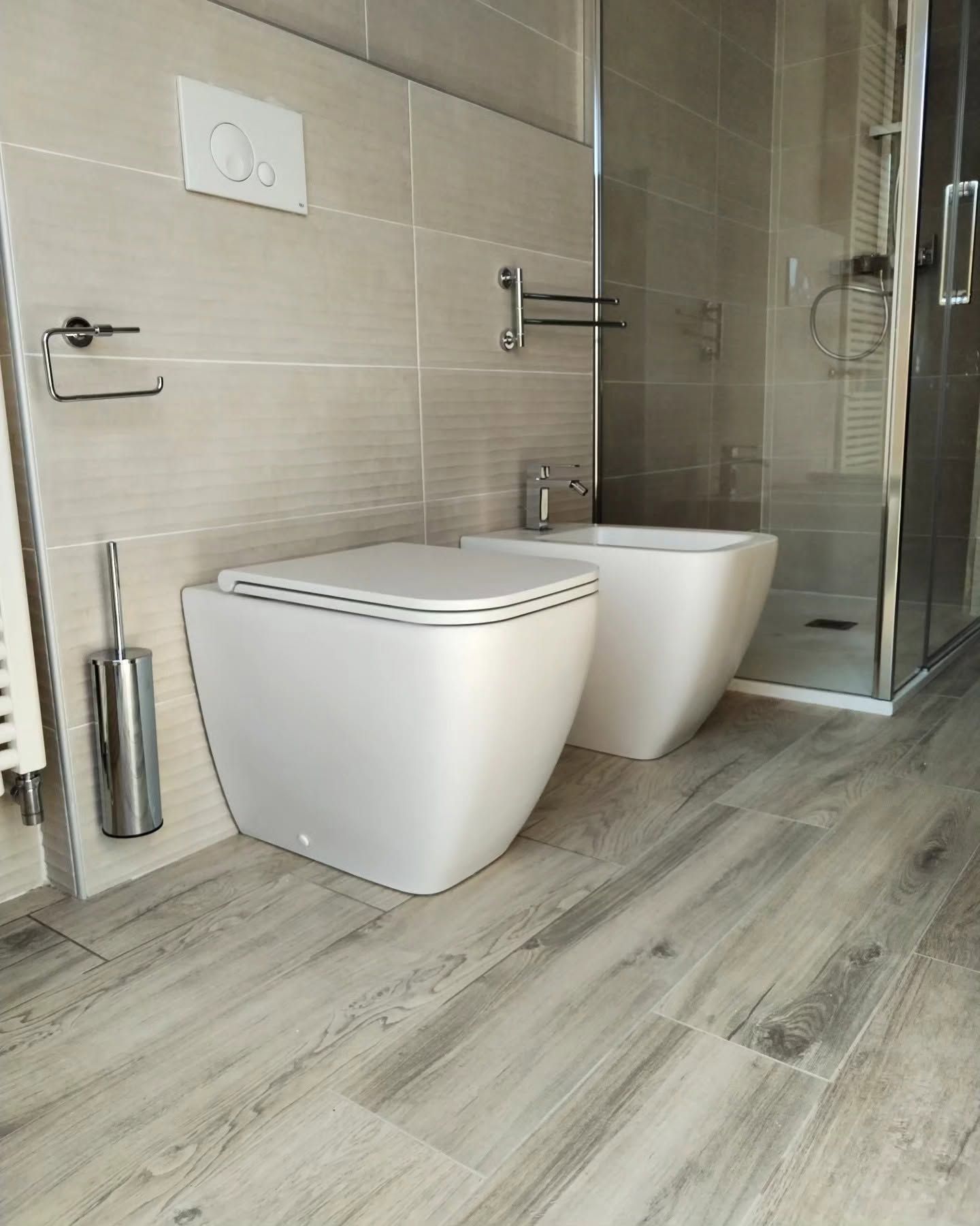 Bagno moderno con due WC sospesi bianchi, doccia in vetro e pavimento piastrellato effetto legno grigio.