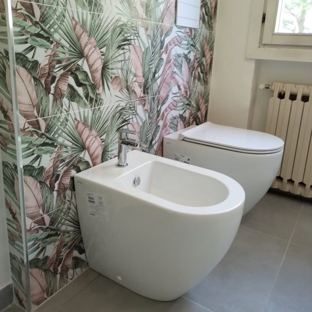 Bagno moderno con bidet e WC bianchi accanto a una carta da parati con motivo a foglie tropicali.
