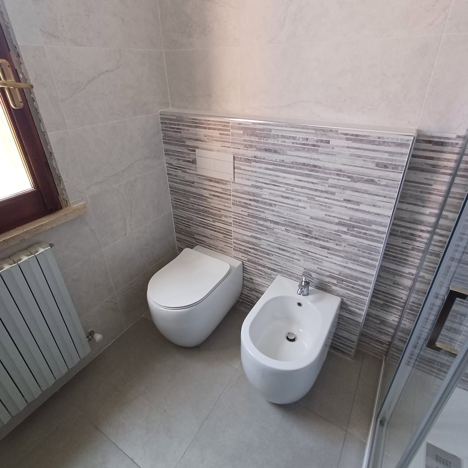Piccolo bagno piastrellato con WC, bidet, finestra e box doccia in vetro.