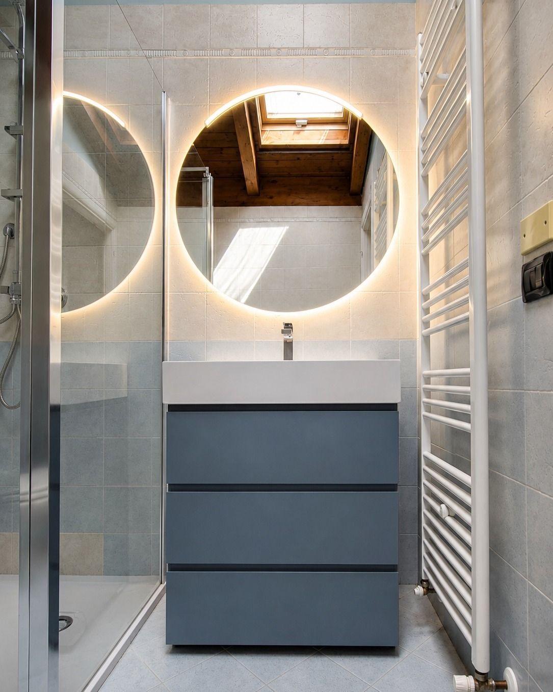 Bagno moderno con specchio rotondo retroilluminato, mobiletto blu, lavabo bianco, box doccia in vetro e termoarredo.