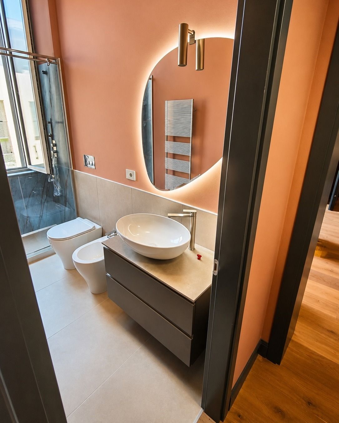 Bagno moderno con specchio rotondo, lavabo a ciotola, WC e doccia in vetro accanto a una parete con dettagli color pesca.