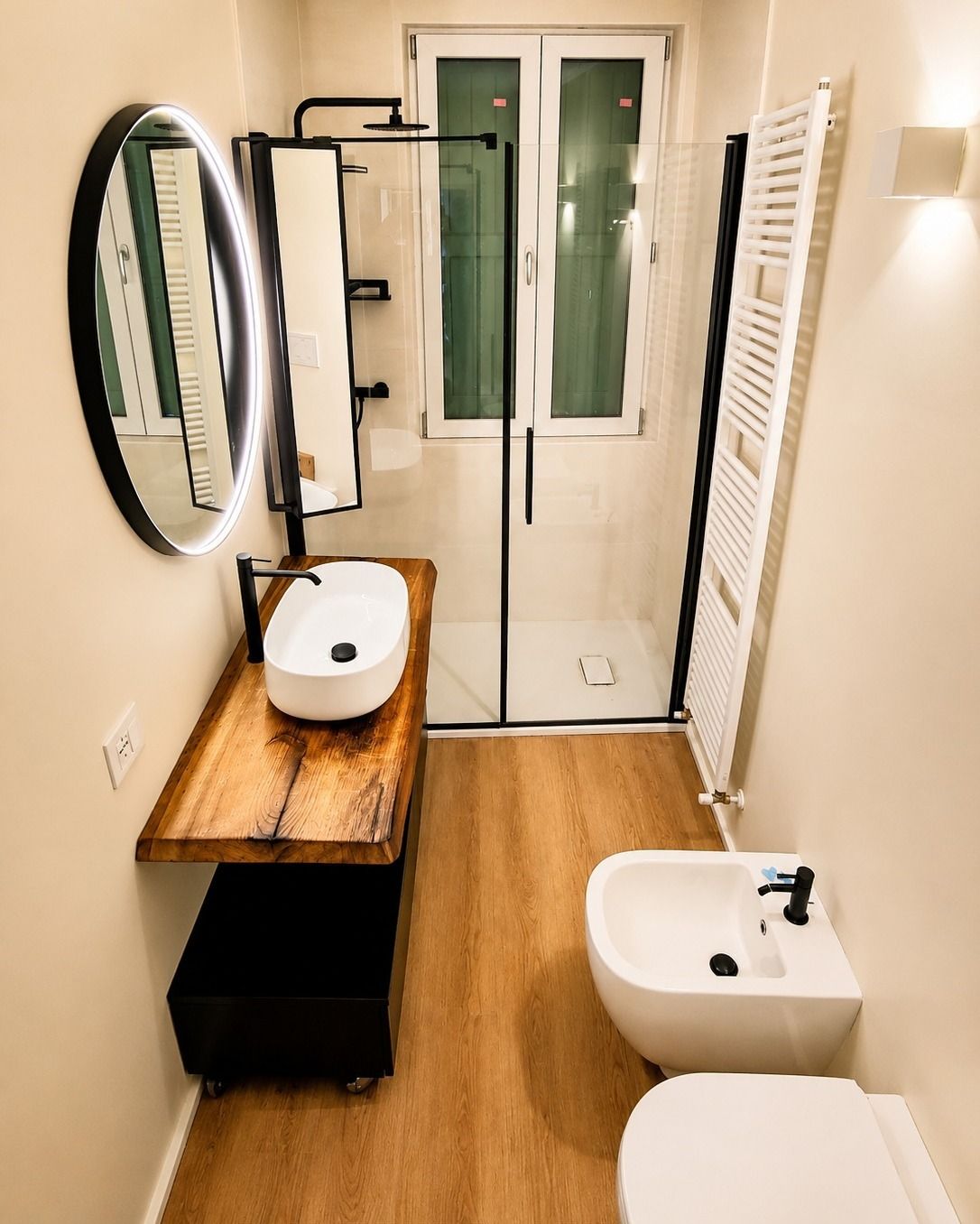 Bagno stretto con lavabo, bidet, specchio, termoarredo e porte doccia in vetro.