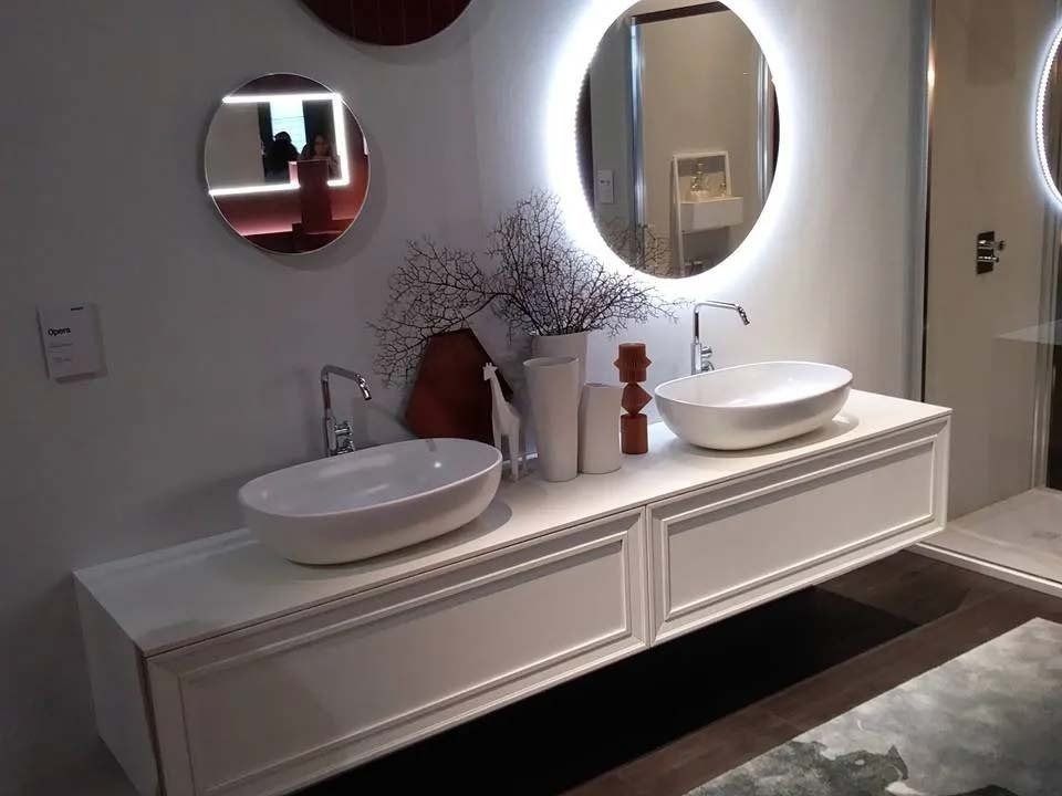 Mobile bagno moderno con due lavabi a ciotola, specchi e composizione floreale, illuminato da luci a parete soffuse.