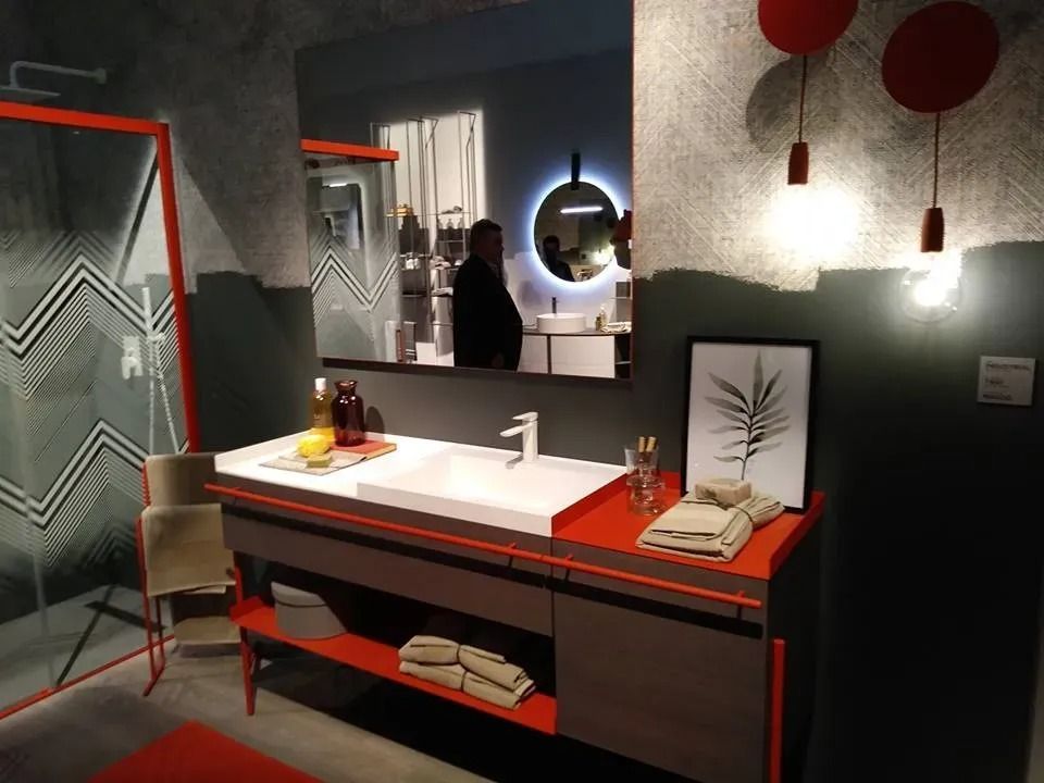 Mobile bagno moderno con piano arancione, specchio rotondo, dettagli rossi e doccia visibile sullo sfondo.