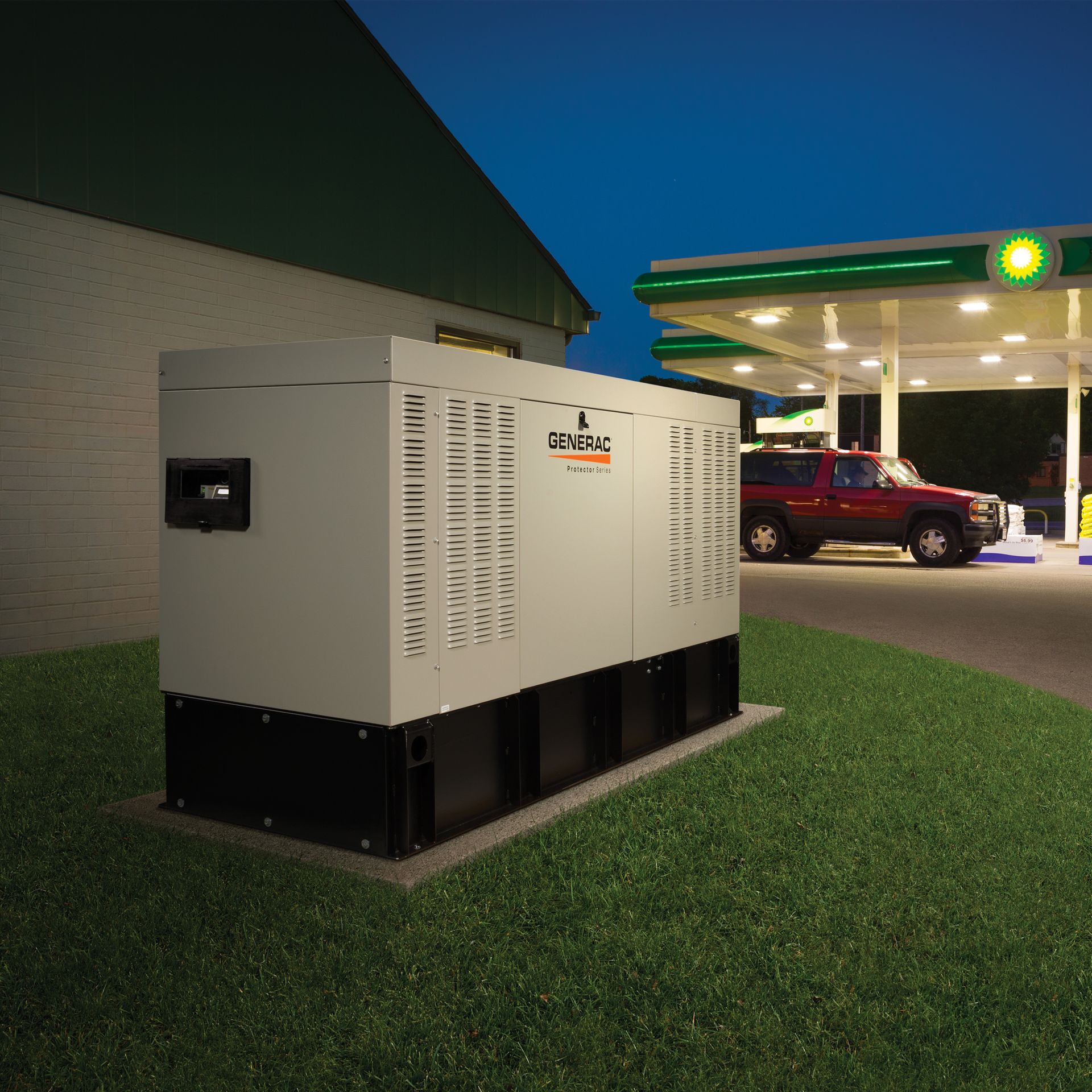 Large, industrial generator on a grassy patch next to a gas station at dusk. A red SUV is at the pumps.