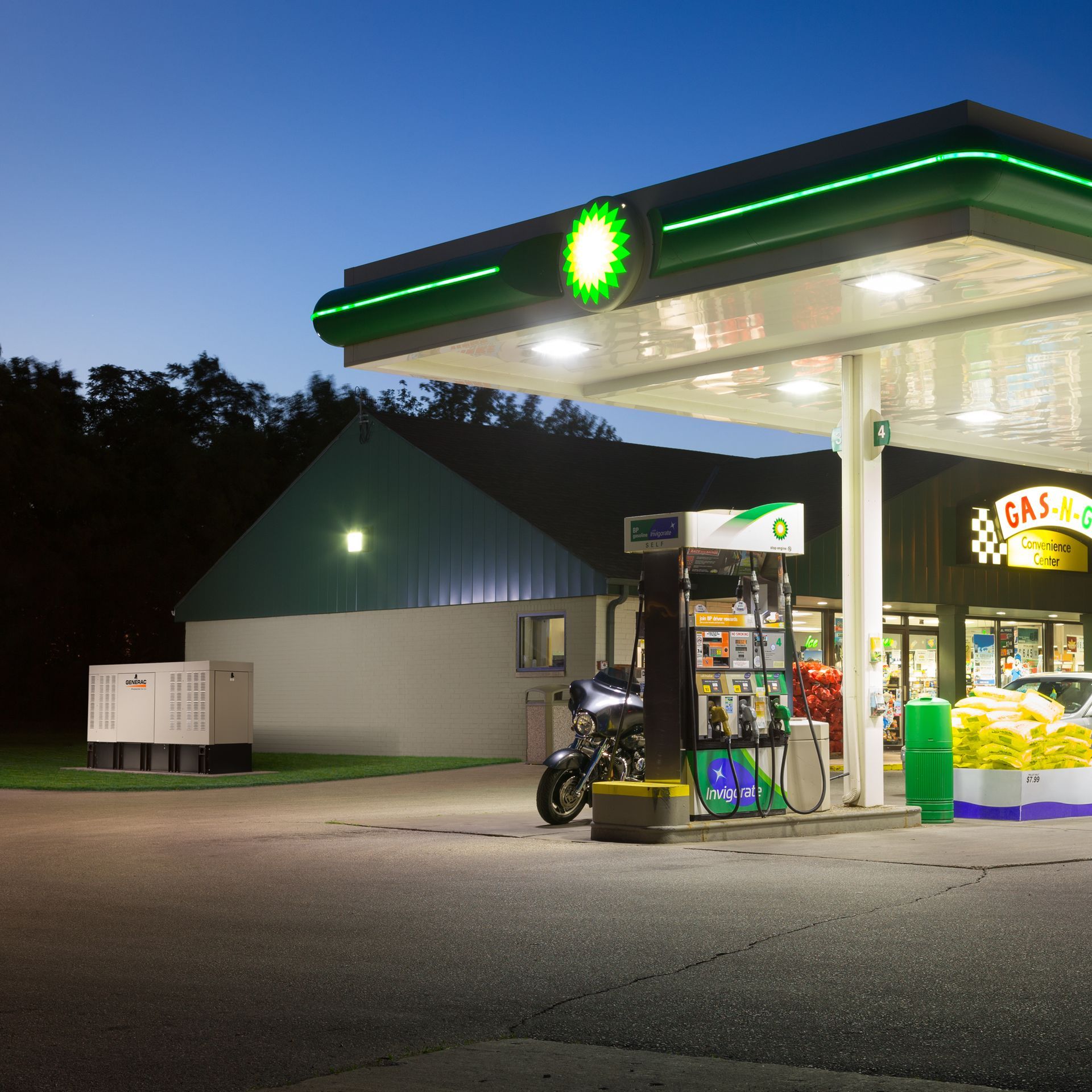 A BP gas station at dusk with a motorcycle parked at a pump and a store visible. The station is lit with green accents.