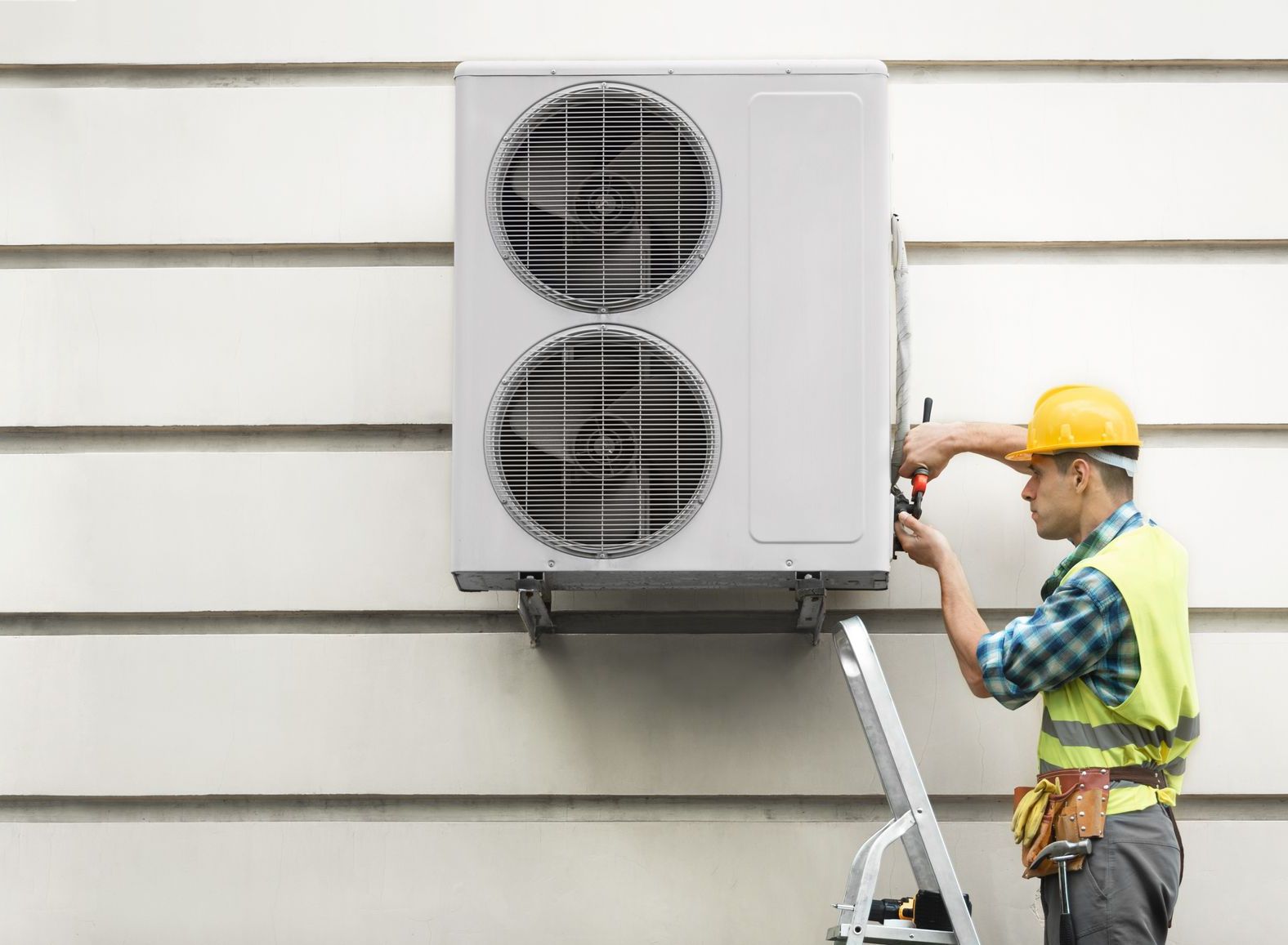 HVAC technician in a yellow vest and hard hat working on an outdoor air conditioning unit mounted on a white wall.