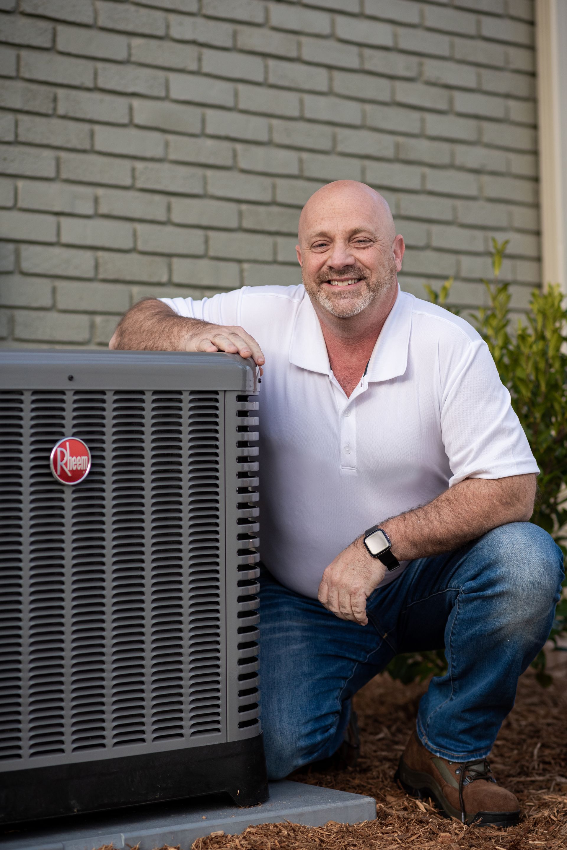 A smiling man in a white shirt and jeans kneels beside a Rheem air conditioning unit. He's outdoors near a brick wall.