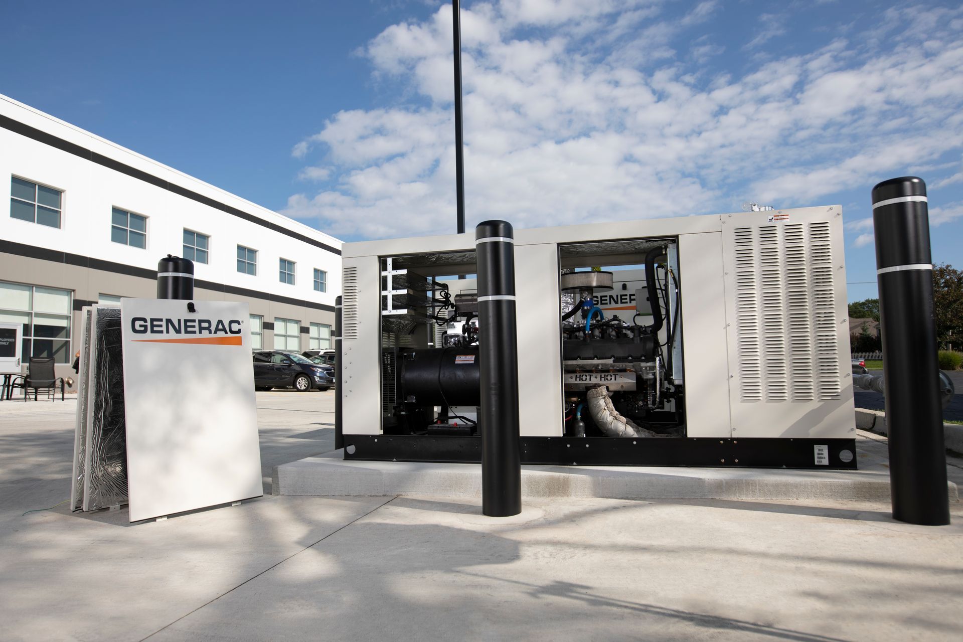 A large, beige Generac generator sits outside a white modern building, protected by black bollards under a blue sky.