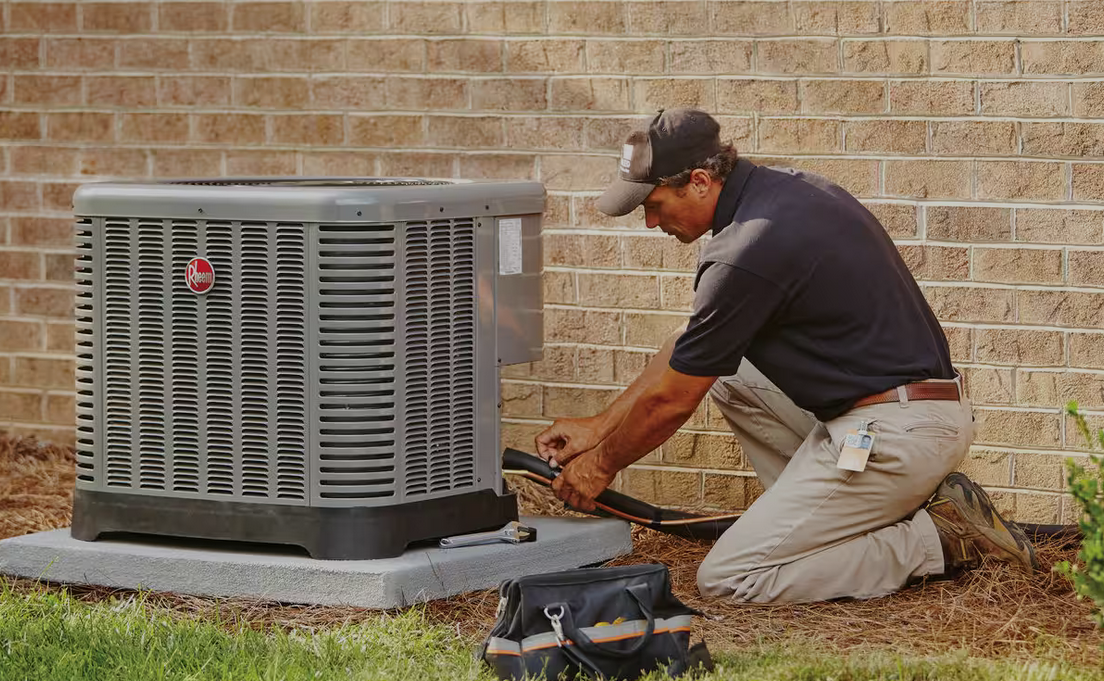 HVAC technician kneeling beside an air conditioning unit, working on it outside a brick building.