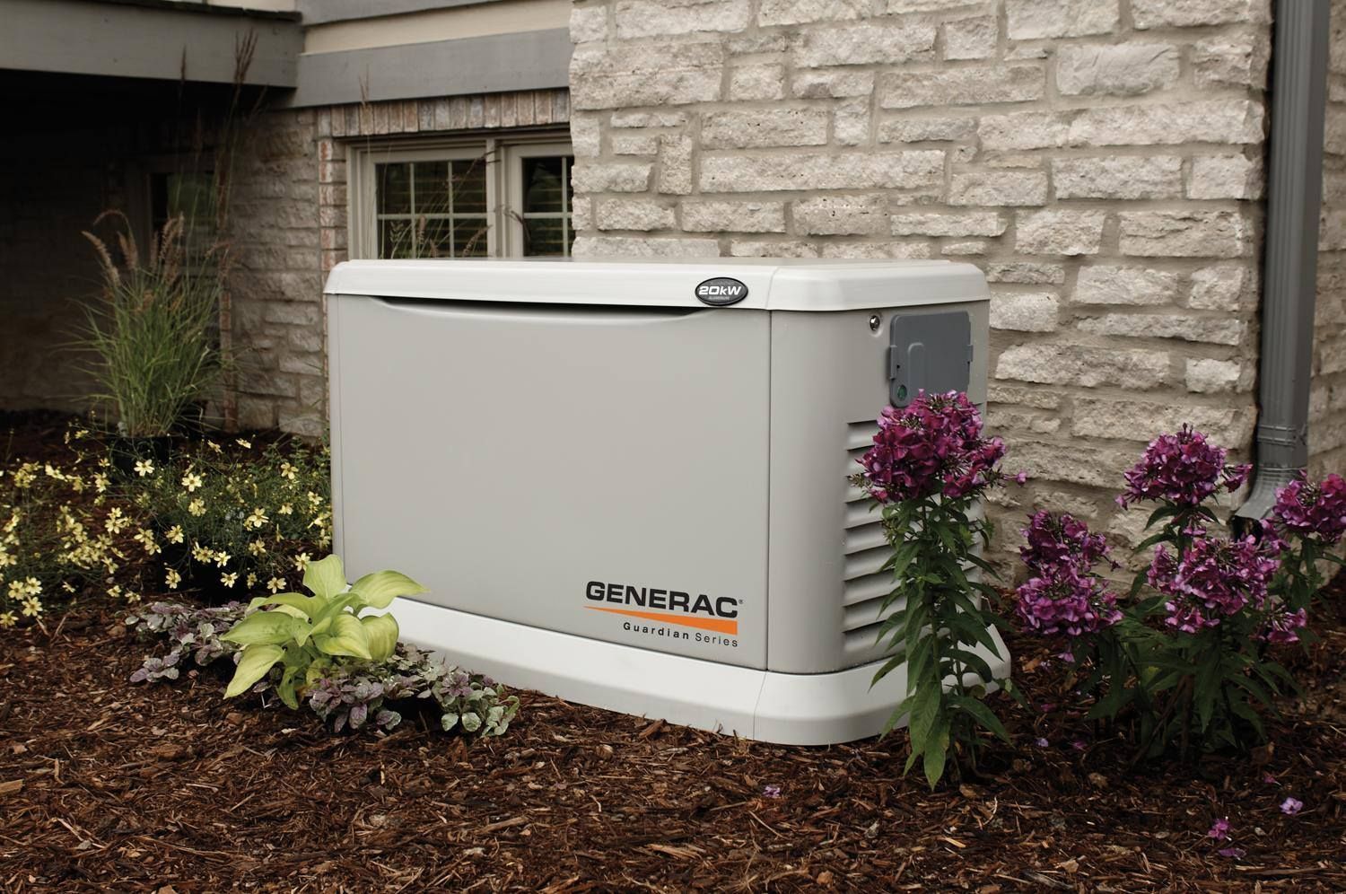 A Generac home standby generator sits on a landscaped base beside a brick building. It is beige.
