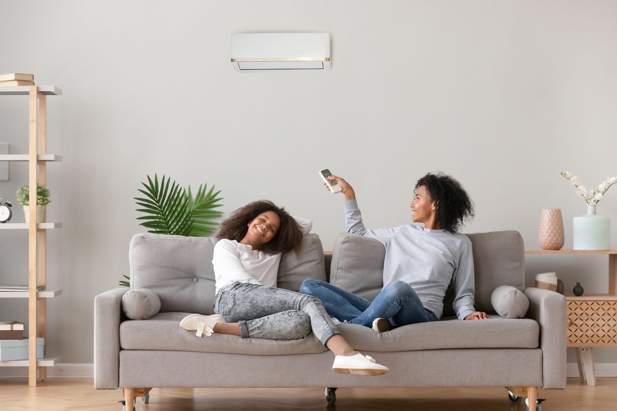 Two people sit on a couch; one holds a remote pointing at an air conditioner. White walls, a simple living room setting.