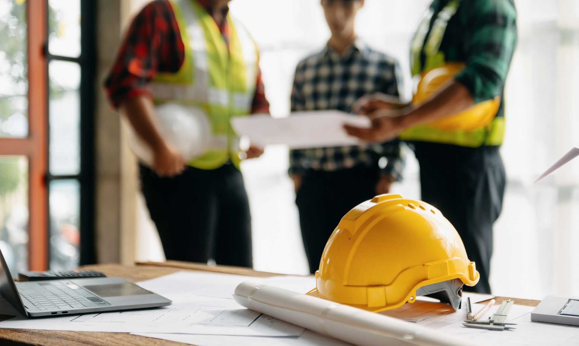 Three construction workers reviewing blueprints, with a yellow hard hat and laptop in the foreground.