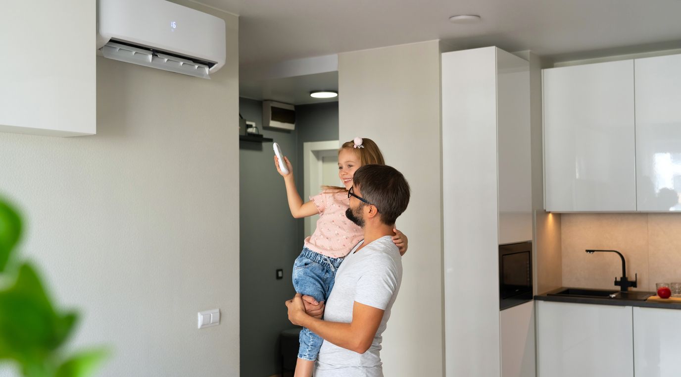 A man holding a girl is using a remote to control a wall-mounted air conditioner in a modern apartment.
